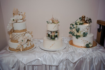 Elegant wedding cake display featuring a three-tiered white cake with gold accents and floral decorations, alongside smaller cakes adorned with similar romantic details, presented on a draped table