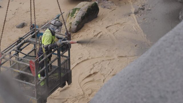 Worker in safety gear uses high-pressure spray to apply concrete on a coastal cliff to prevent erosion and collapse