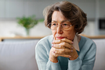 Thoughtful sad elderly woman sitting on couch, pensive older female looking into distance, thinking...