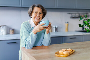 Cheerful senior woman in good mood drinking tea on kitchen, looking and smiling at camera. Rest,...