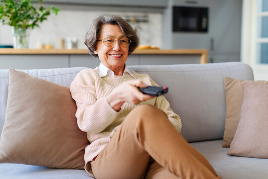 Happy smiling senior woman sitting on couch in the living room, pointing tv remote controller to switch channels, watching favorite movie online series