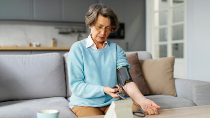 Serious senior woman using medical device to check her blood pressure measurement, suffering from hypertension, sitting on couch at home