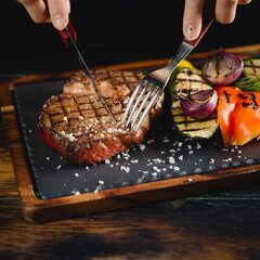 Woman eating grilled ribeye steak served on a dark slate plate with vegetables, scene with cutlery - fork and knife