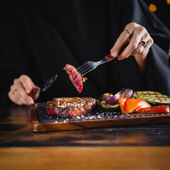 Woman eating grilled ribeye steak served on a dark slate plate with vegetables, scene with cutlery - fork and knife