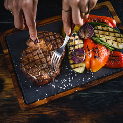 Woman eating grilled ribeye steak served on a dark slate plate with vegetables, scene with cutlery - fork and knife