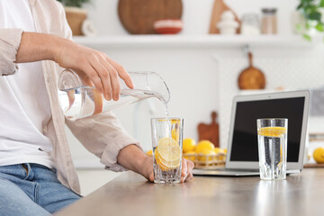 Young man pouring water into glass with lemons at table in kitchen, closeup
