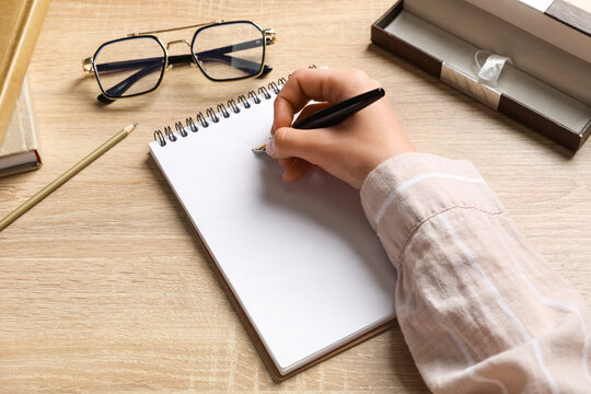 Woman writing something in notebook, pencil and eyeglasses on wooden background, closeup