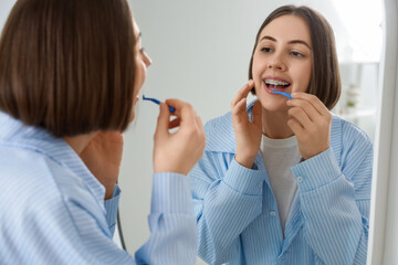 Young woman with dental braces cleaning teeth near mirror in bathroom