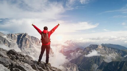 A triumphant hiker raises both arms atop a rocky peak above swirling clouds. Concept of adventure success and personal achievement.