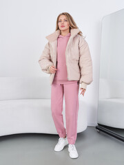 Young woman in pink tracksuit with matching puffer jacket stands by a mirror. She wears white sneakers and has long blonde hair. The decor includes a white couch and a simplistic setting © Dmitry Tsvetkov
