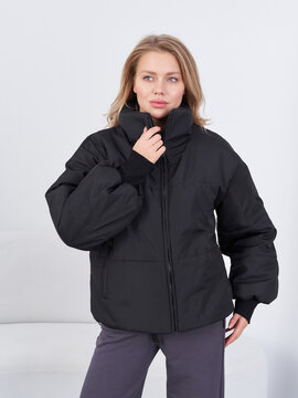 Female model in a black puffer jacket with a high collar, adjusting the collar with her right hand. Her expression is confident, eyes looking slightly to the side