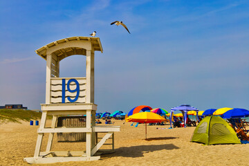 A landscape of a white lifeguard tower on a sandy beach with a seagull in flight and colorful umbrellas at the Carolina Beach in North Carolina on a sunny summer day.