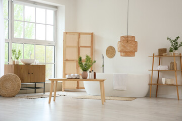 Interior of stylish bathroom with bathtub, folding screen, commode and green blossoming branches in...