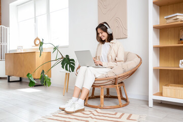 Young woman with headphones using laptop in cocoon chair at home