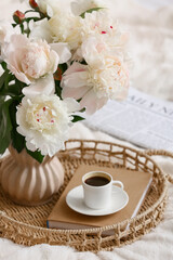 Tray with peony flowers in vase, cup of coffee and book on bed, closeup