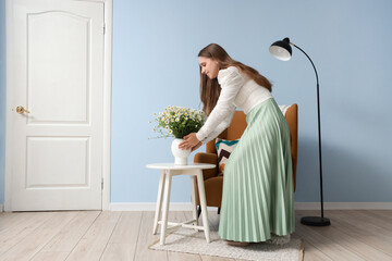 Young woman putting vase with chamomiles on table at home