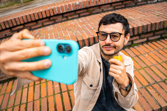 A man with a beard and glasses taking a selfie while eating a peach he is holding outdoors. Concept of peach month, healthy food, wellness, and technology. High quality photo