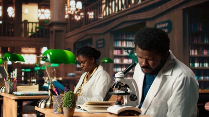 Male student researching viruses during a microscope analysis and academic reading in a classic library. Med school pupil reflects dedication to biology and exam preparations. Camera A.