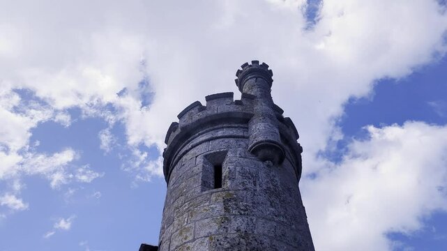 A low-angle shot looking up at a stone tower of Monterreal Castle in Baiona, Galicia, Spain. Fast-moving clouds pass over the medieval fortress battlements against a vivid blue sky.