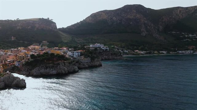 Aerial drone footage of Porticello Sicily showing vibrant cliffside homes above the sea with Monte Catalfano and the Tyrrhenian coastline toward Bagheria and Aspra in soft afternoon light