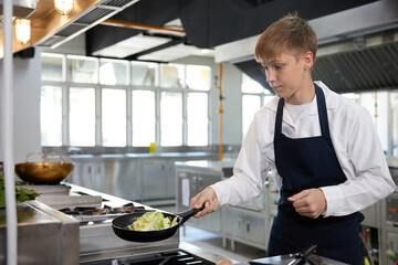 young chef cooking stir fried vegetables with spatula and pan in the kitchen