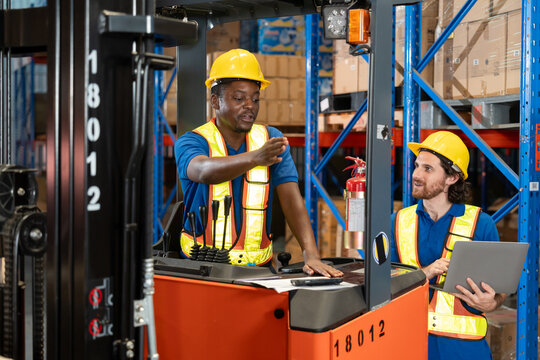 Black male warehouse worker standing at forklift, pointing forward while explaining procedure to bearded colleague holding laptop. Both wearing helmet and vest inside organized storage facility.