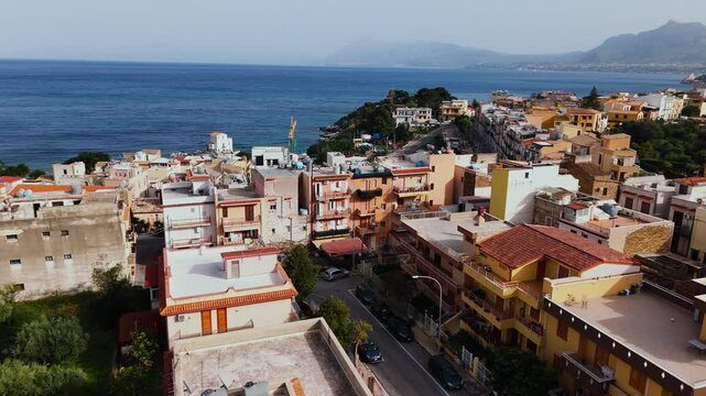 Slow motion drone flight above colorful buildings and narrow roads in Porticello, Sicily, revealing the Tyrrhenian Sea and distant Palermo coastline in golden daylight