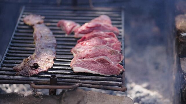 Close up of traditional Argentine asado with skirt steak (entra&ntilde;a) and top sirloin cap (picaña) cooking on metal grill over wood embers, static real time shot, rustic barbecue scene, natural daylight