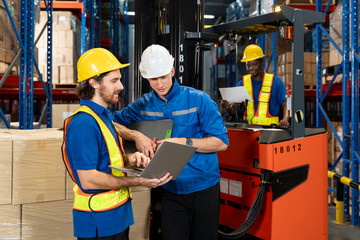 Three male warehouse staff from diverse backgrounds in uniform and helmet collaborating using laptop and documents. Two discussing at front, one on forklift reviewing paper inside busy storage 