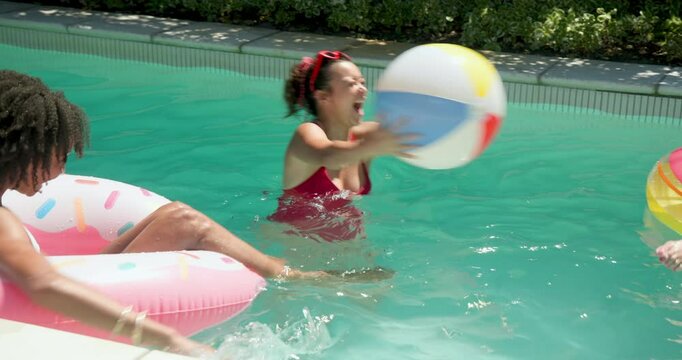 Laughing Diverse female friends enjoying pool day, playing with beach ball and relaxing on float