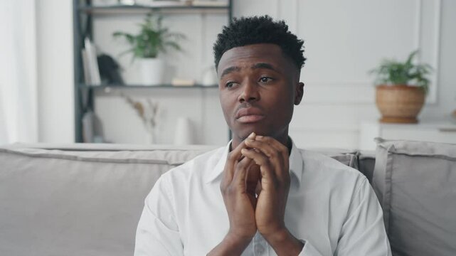 Emotional portrait of young black man sitting alone in living room and thinking. Medium shot indoor with handsome african guy in white shirt, worried single male person dreaming about future, feel sad