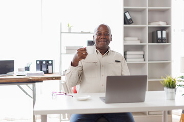 Happy smiling senior man using Laptop while holding coffee cup sitting at his cozy workplace office.
