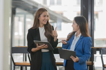 Two Asian businesswomen holding tablets and laptops, standing in a meeting discussing management in an office.