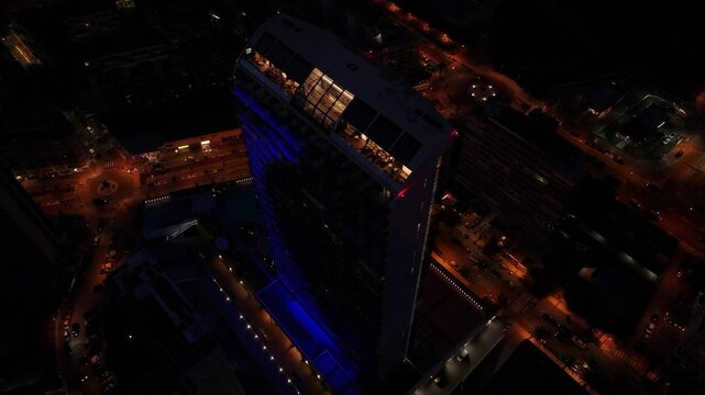 Nighttime drone orbiting above the Estimar Calpe Hotel in Calp, Spain, showing the building&rsquo;s blue lighting, rooftop, and surrounding city streets and modern architecture