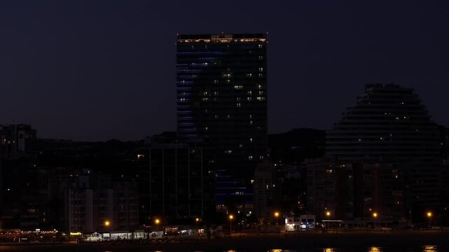 Night drone zoom-in on Estimar Hotel, Calp, Alicante, Spain, showing lit skyscraper, urban buildings, and distant mountains, city lights glowing below, clear night sky, iconic coastal skyline.