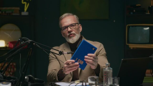 Stab medium POV shot of mature Caucasian male author talking in microphone about bestselling book and looking at camera in podcast studio