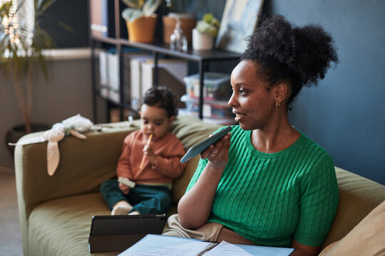 African American woman working from home while toddler plays in background. Cozy setting with plants and bookshelves creating a warm and productive environment - Powered by Adobe