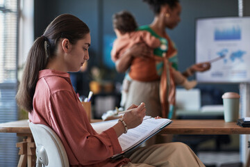 Employees working on various tasks in modern office environment, with focus on one person writing...