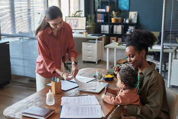 Mother holding child while working from home, discussing documents with female colleague at office desk, multitasking and balancing work and family life in cozy home office setting