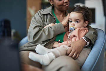 African American mom cuddling baby with pacifier while sitting on couch. Warm scene showing bonding...