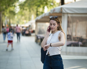 Busy young woman multitasking on phone call and checking time outdoors
