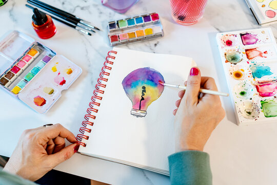Woman painting light bulb with watercolors on spiral notebook at table
