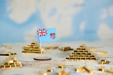 Fiji's national flag stands prominently among numerous gold bar piles on a blurred world map,...