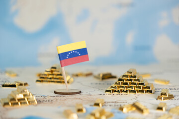Venezuelan flag miniature standing amidst numerous gleaming gold bars on a blurred global map,...