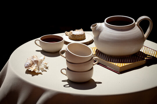 Ceramic teapot and cups on a table with a shell and book in sunlight