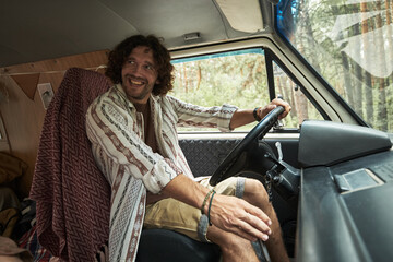 Man with curly hair is sitting in drivers seat of a van, driving through a forested area, wearing a striped shirt and shorts, radiating happiness and relaxation