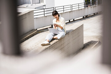 Teenage girl with laptop using smart phone on retaining wall during sunny day