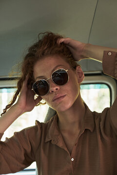 Woman sitting inside vehicle wearing sunglasses and brown shirt, leaning back and resting hands on head, basking in casual and relaxed atmosphere