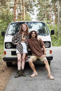Young couple standing and sitting in front of a vintage van in a wooded area, sharing a joyful moment surrounded by tall trees