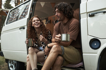 Couple enjoying outdoors by camper van, holding mugs, and laughing in forest setting, capturing moment of relaxation and fun during camping trip